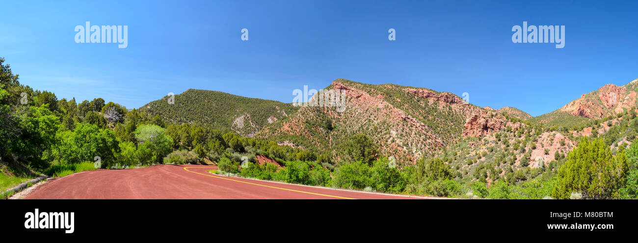 Vista panoramica di kolob canyon su pull-off. Verdi alberi e cespugli, rosso strada asfaltata, cielo blu con rosso e arancio montagne e colline. Foto Stock
