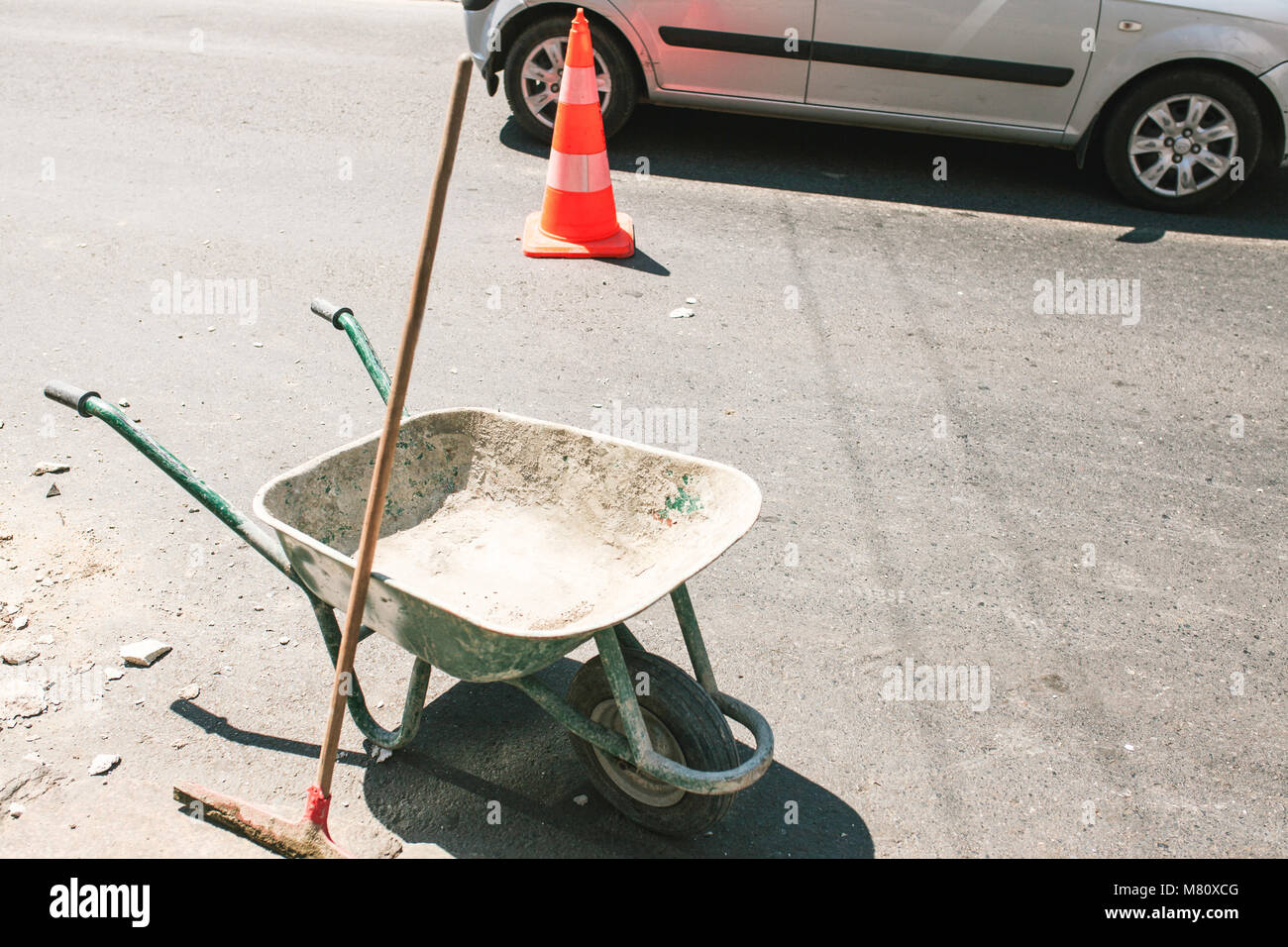 Immagine di rustica costruzione sporco carriola con push ginestra sulla strada, arancione per la costruzione di strade di cono in background. Foto Stock