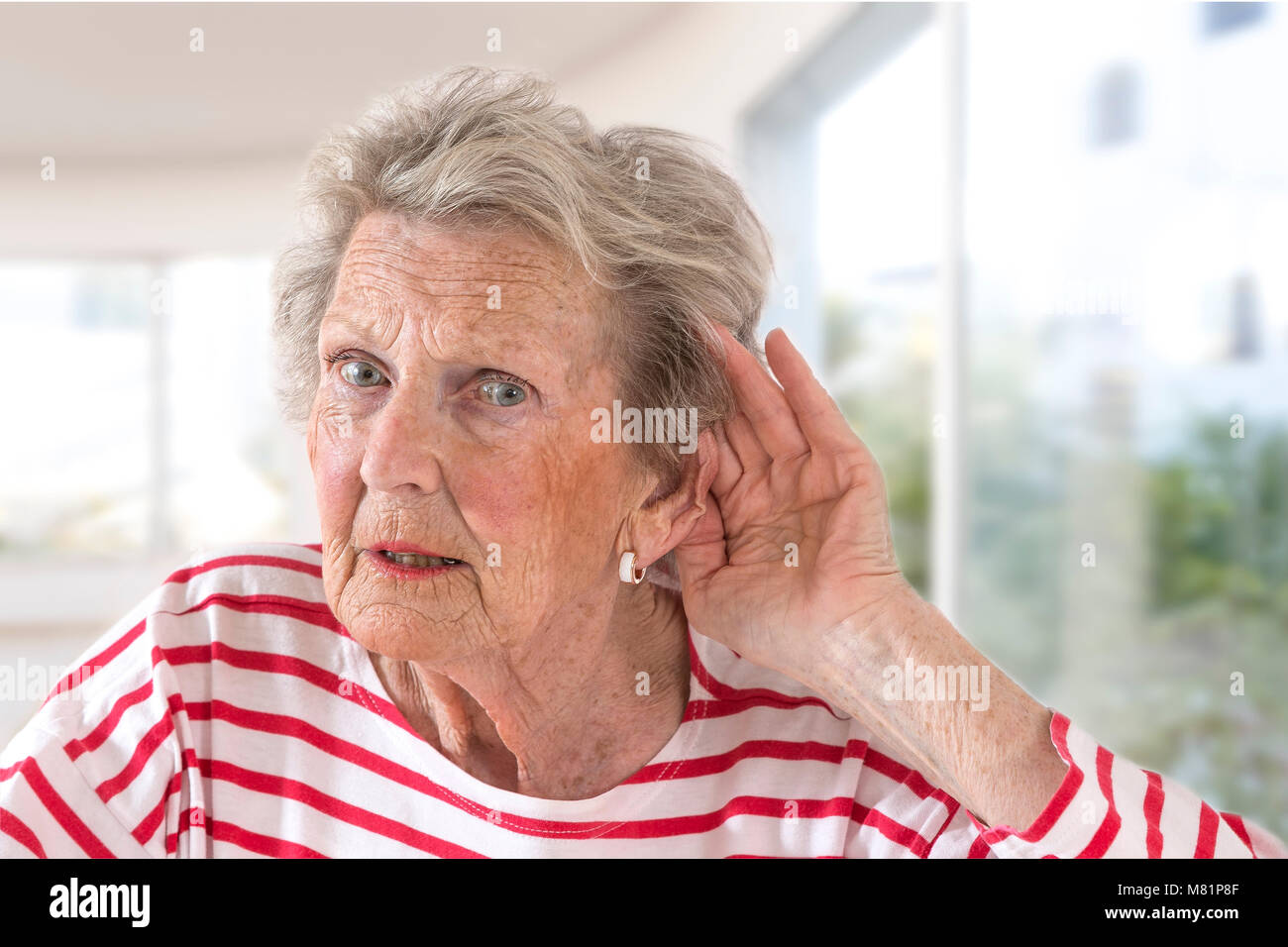 Signora anziana con problemi di udito a causa dell'invecchiamento tenendo la mano al suo orecchio come lei le lotte per sentire, vista di profilo su un grande sfondo di Windows Foto Stock