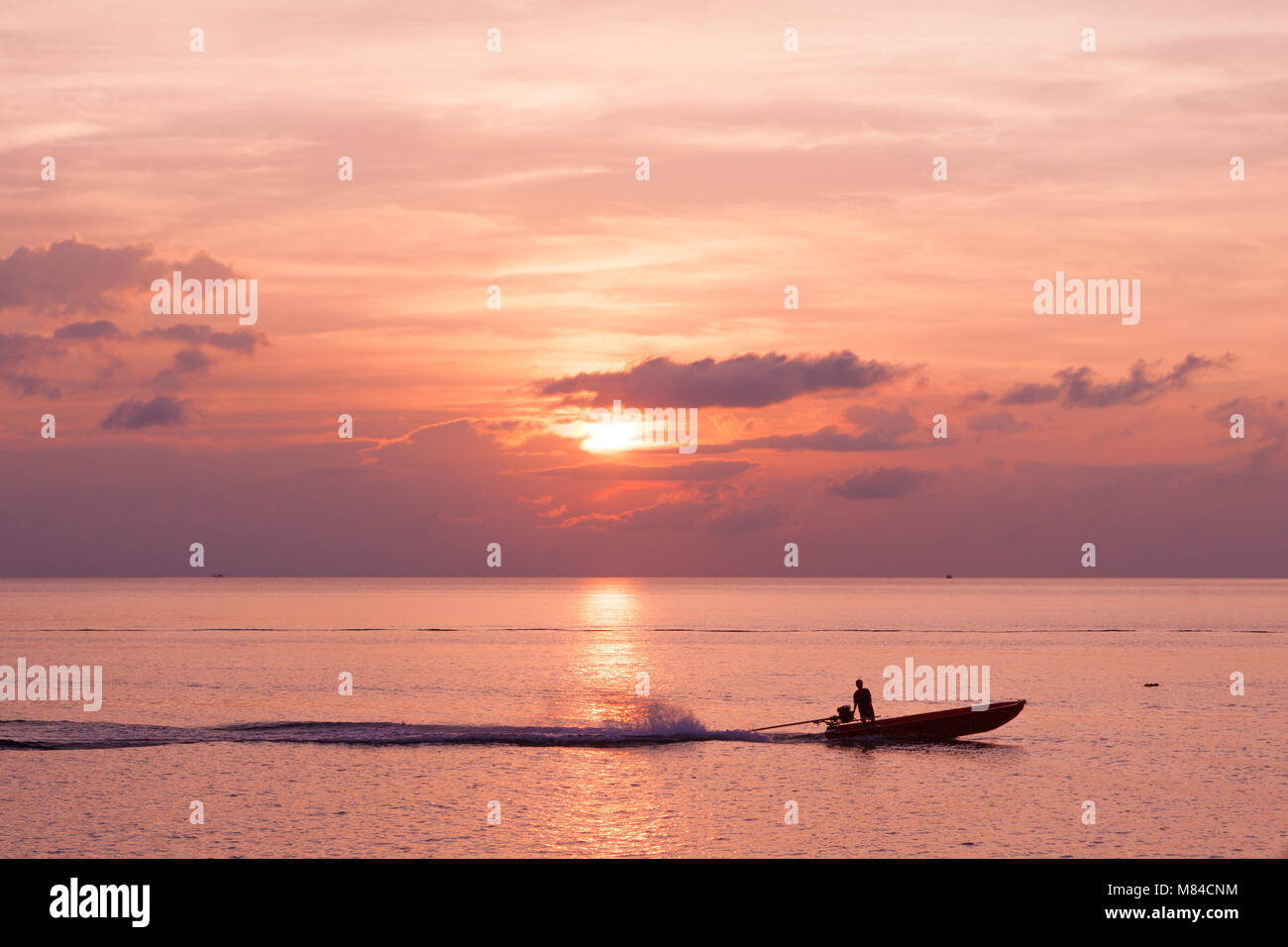 Silhouette di un uomo su una barca, lasciando una scia nel mare sotto una rosa NUVOLOSO TRAMONTO Foto Stock