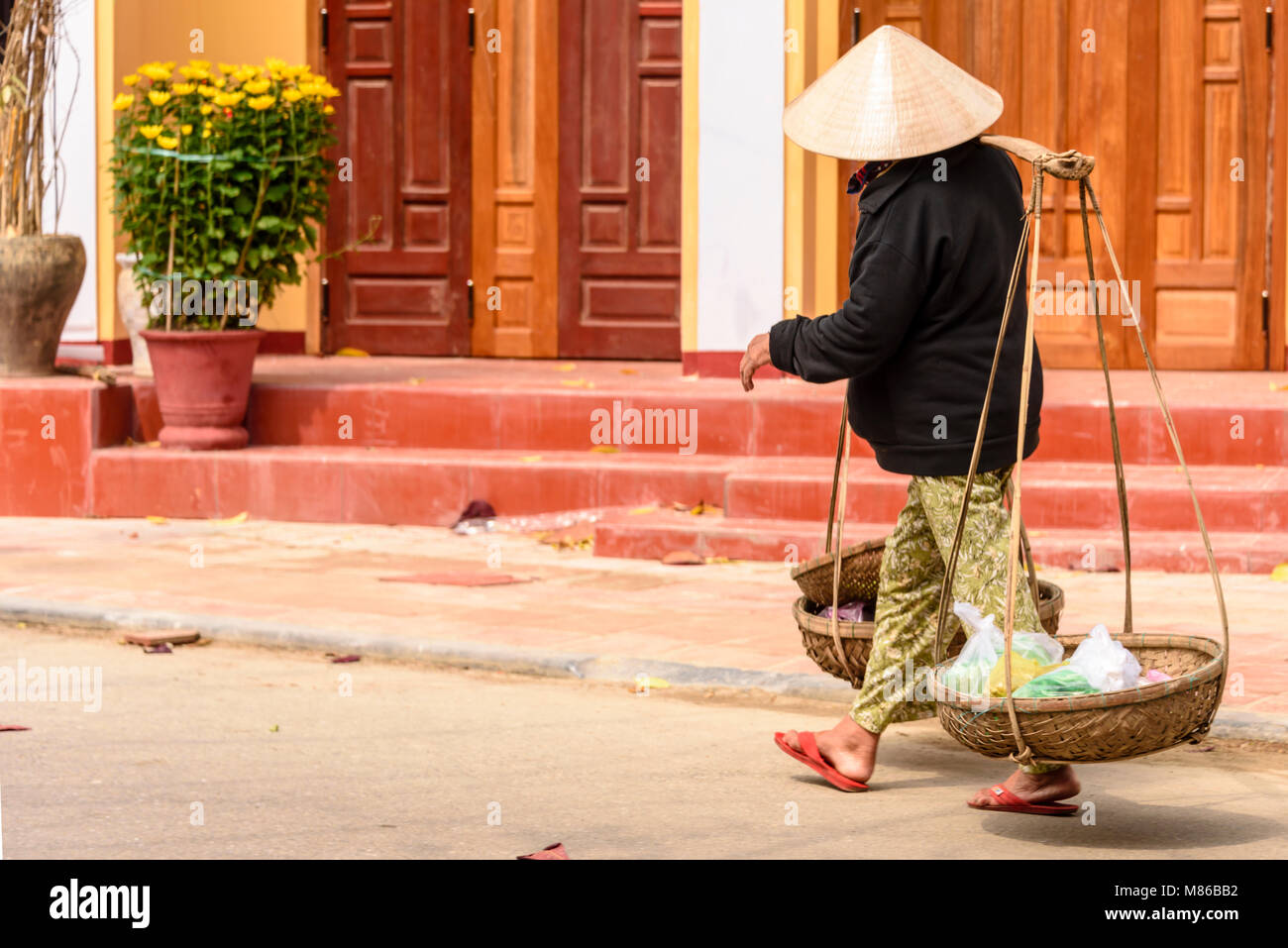 Una donna che indossa un tradizionale di bambù cappello conico porta alcuni elementi nei cestelli appesi da lei bambù Palo portante. Foto Stock