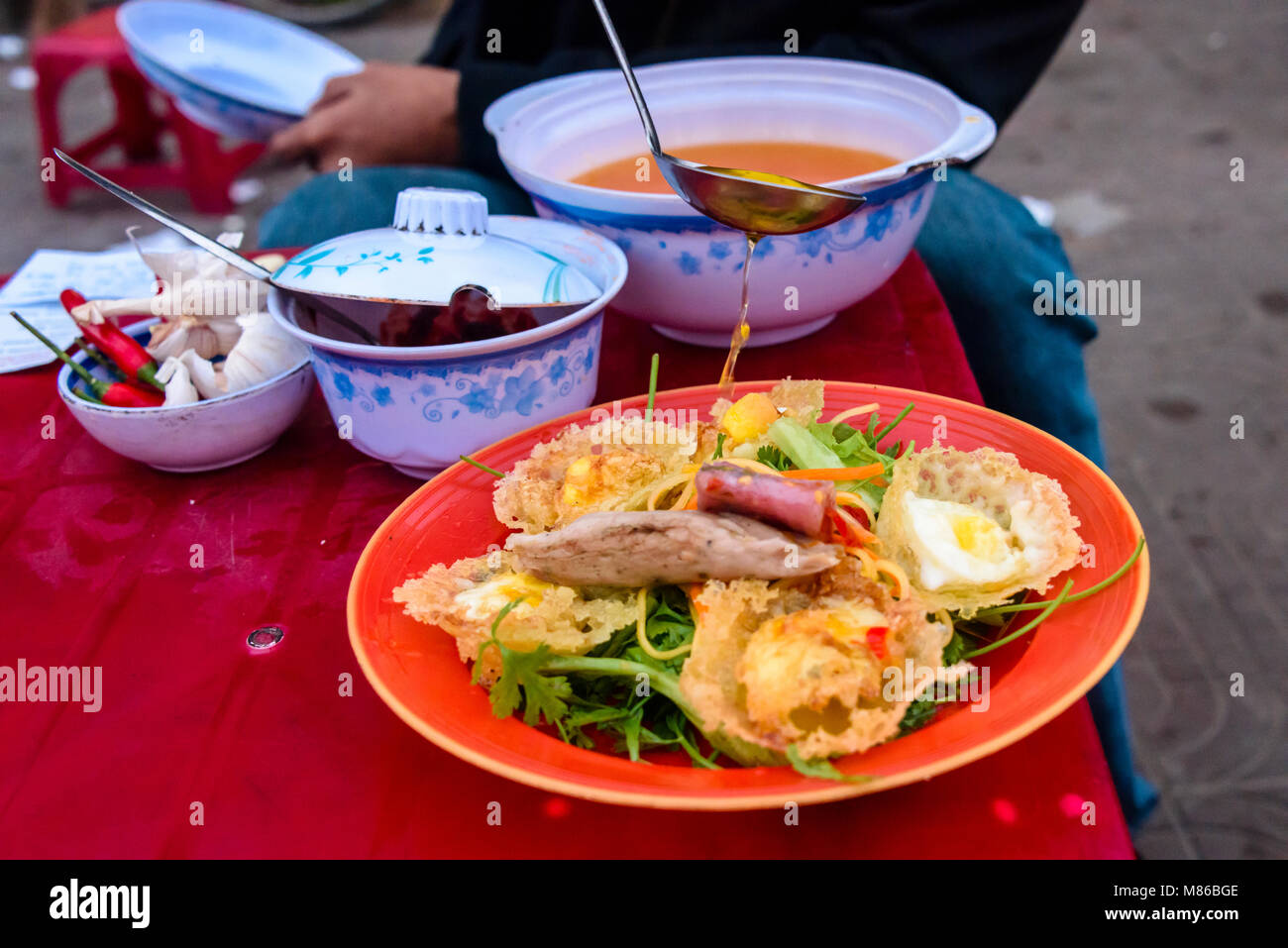 Cucina di strada, consistente di frittelle di riso con uova di quaglia, carne di maiale e manzo, su un letto di foglie verdi, ha un miele e salsa chili versato su di esso, in Hoi An, Vietnam Foto Stock