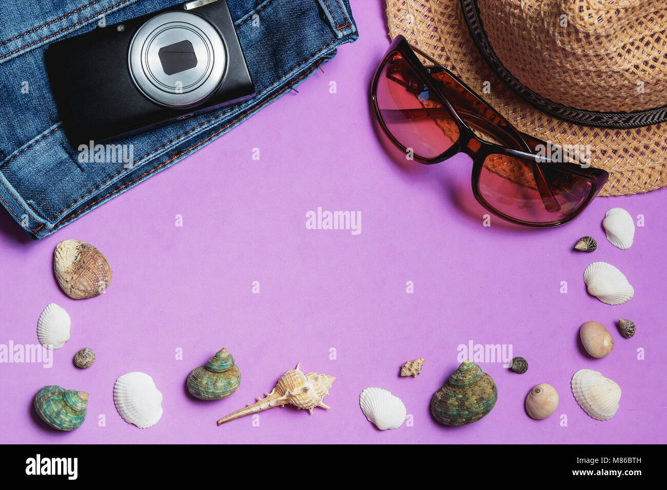 Jeans, occhiali da sole, Fotocamera, marrone cappello, conchiglie su sfondo lilla. Vista dall'alto concetto di viaggio con Copyspace. Foto Stock