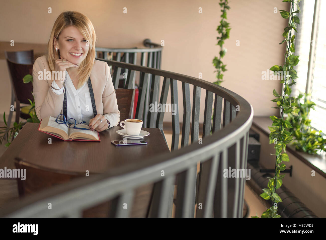 Sorridente giovane donna bionda con gli occhiali in un ristorante a leggere un libro e bere un caffè durante una pausa di lavoro. Pausa caffè. Tempo rilassante. Azienda Foto Stock