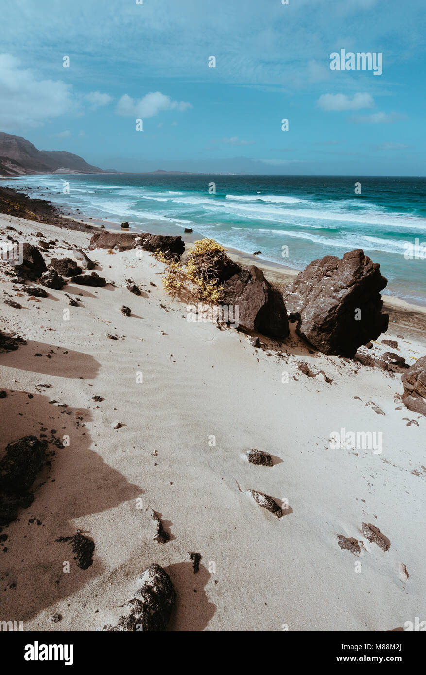 Enorme pietra vulcanica macigno sulle spettacolari dune di sabbia bianca di remote deserta spiaggia Praia Grande. Ocean colpire coste in background. Vegetazione arida. Calhau, Sao Vicente Isola Capo Verde Foto Stock