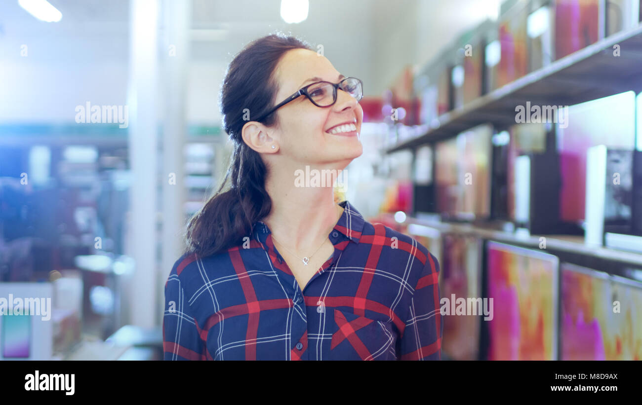 Bella giovane donna cammina nel negozio di elettronica di ripiani di esplorazione con i modelli più recenti di 4K UHD TV. Sorride in quel grande e luminoso negozio moderno Foto Stock