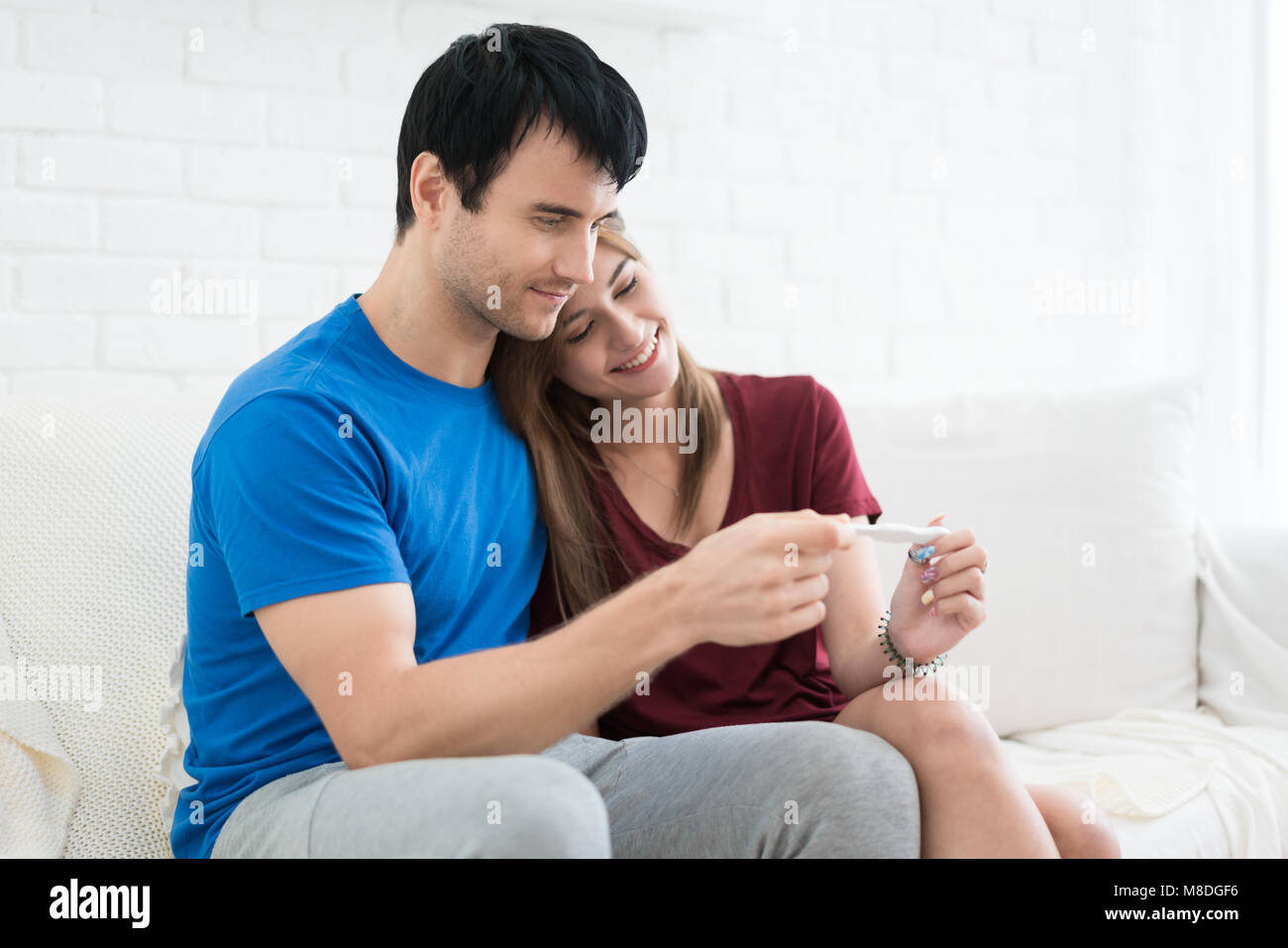Primo piano della felice giovane donna che comprenda l'uomo dopo positivo al test di gravidanza seduta oltre il marito in camera Foto Stock
