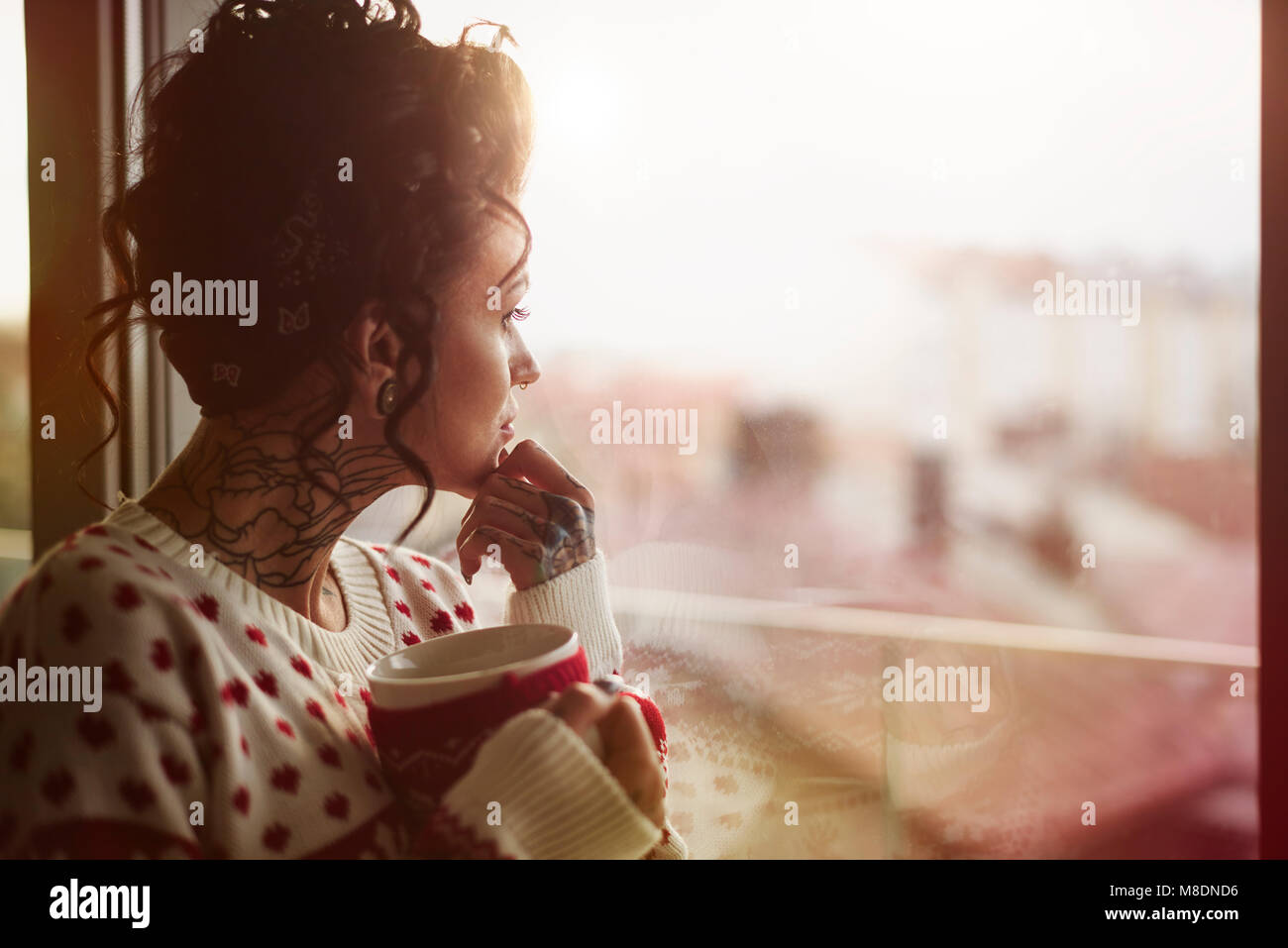Giovane donna guardando fuori della finestra, tenendo una bevanda calda, espressione pensosa Foto Stock
