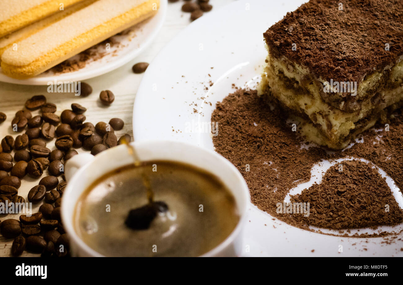 Il tiramisù dolce italiano con cuore di cioccolato grattugiato sulla piastra bianca con tazza di caffè splash scende, biscotti savoiardi e sparse chicco di caffè Foto Stock