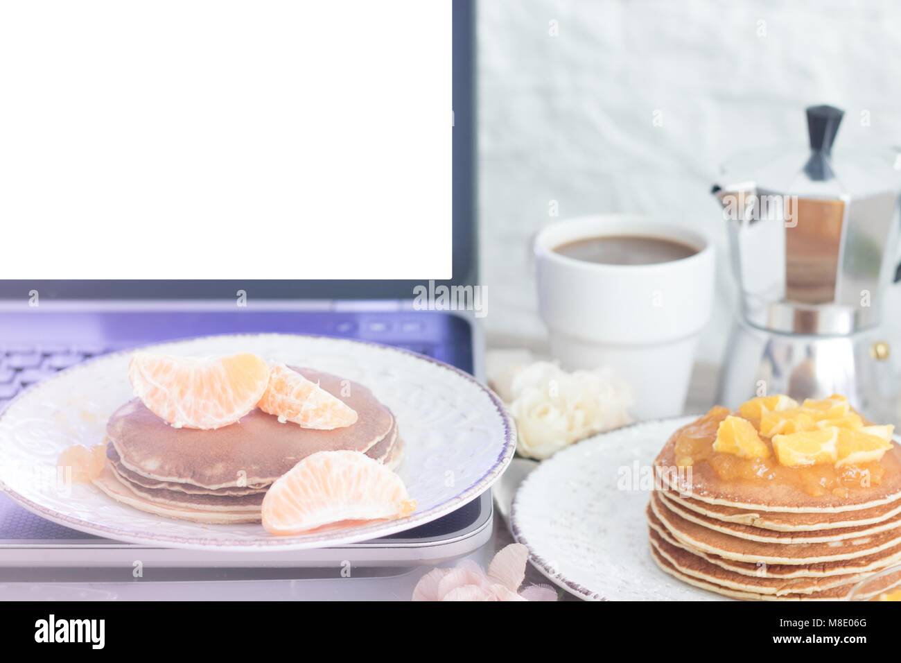 Computer portatile con il bianco schermata vuota sul tavolo con prima colazione: Frittelle con spicchi di mandarini e orange marmellata e caffè con metallo POT del caffè. Vista frontale. C Foto Stock