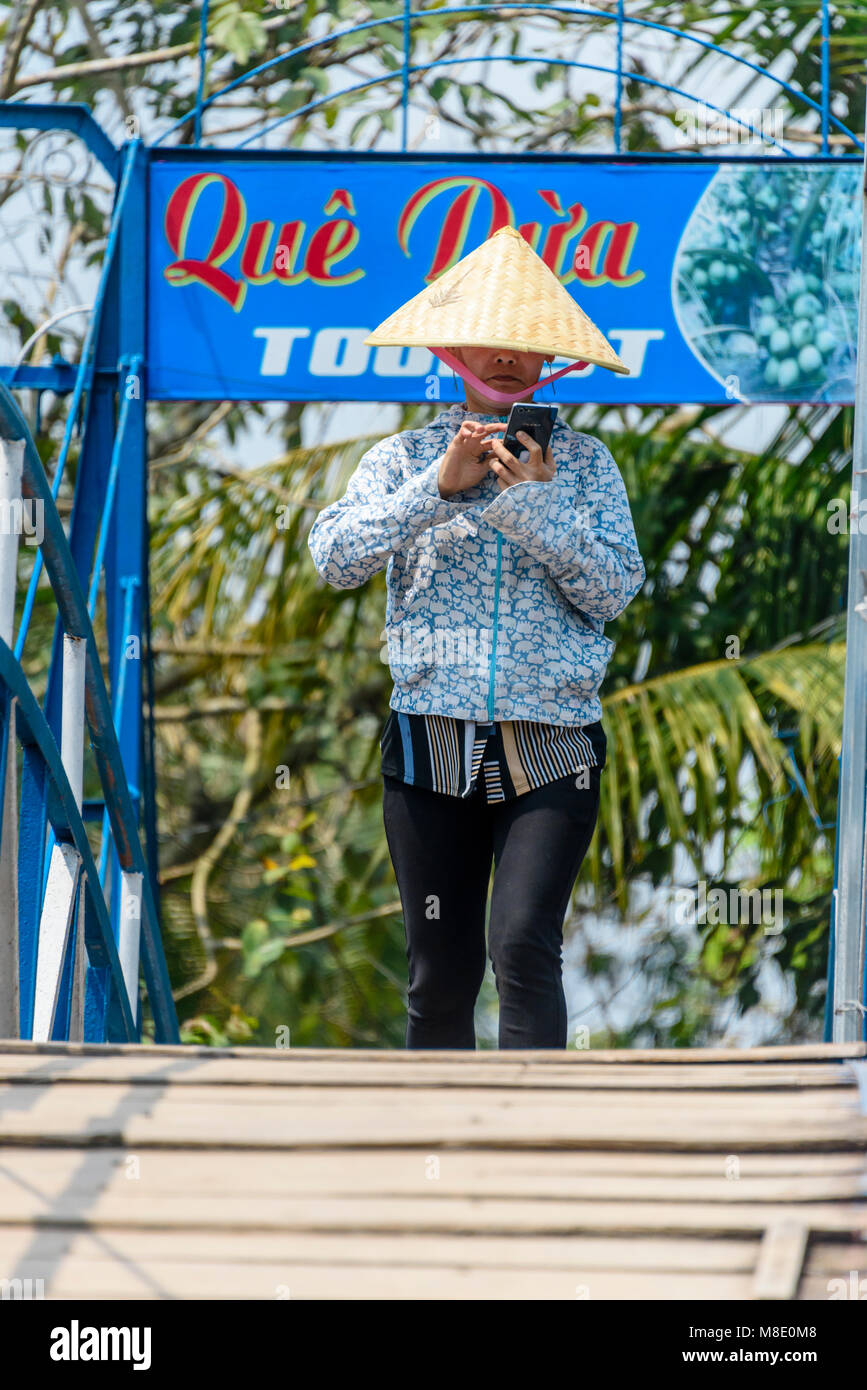 Una donna che indossa un tradizionale vietnamita conico di bambù hat controlla il suo smartphone come lei cammina su un ponte, Vietnam Foto Stock