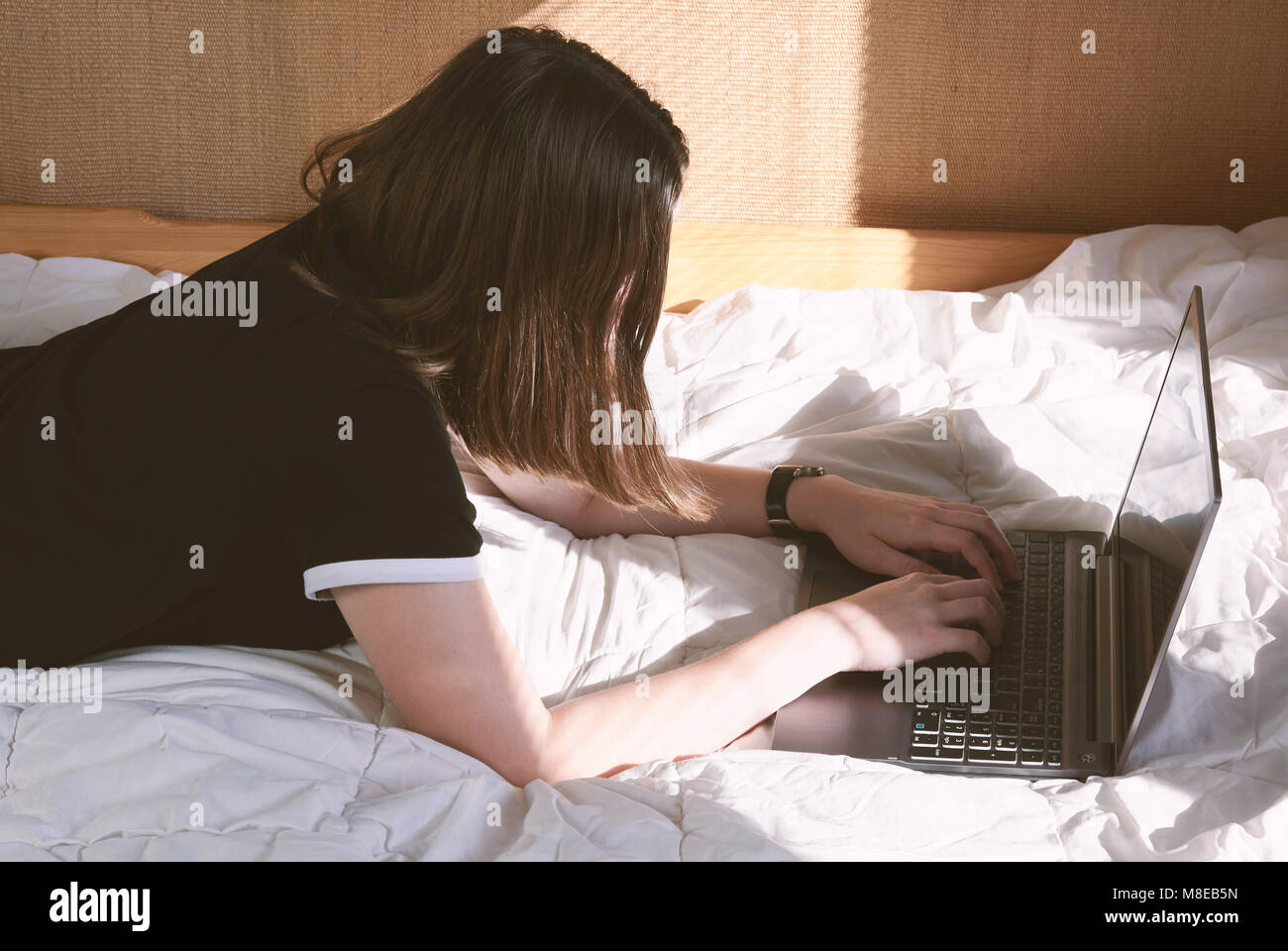 Ragazza giovane giacente sul letto lavorando su laptop - mattina con il sole Foto Stock
