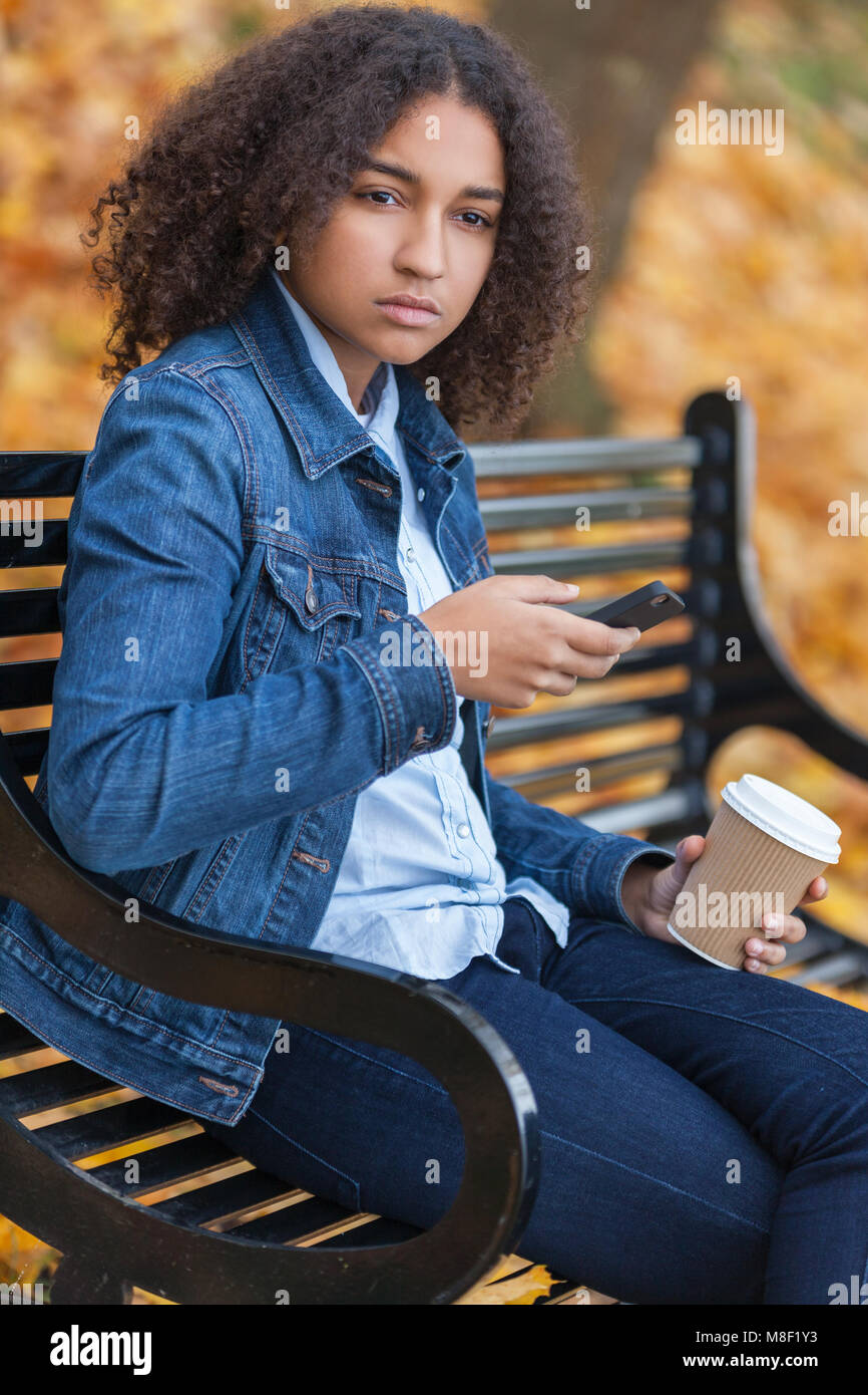 Bellissima gara di misto americano africano ragazza adolescente femmine giovane donna che fa uso del telefono cellulare e di bere il caffè da asporto al di fuori seduta su una panchina nel parco i Foto Stock