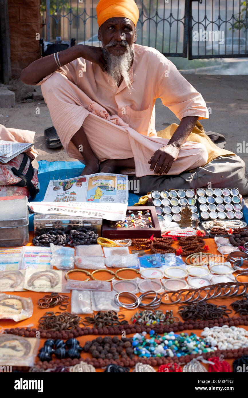 Sadhu indiano - santo uomo seduto sul marciapiede e la vendita di articoli di gioielleria e mala talloni sulle strade di Jodhpur in India Rajasthan. Egli è vestito di luce rosa Foto Stock