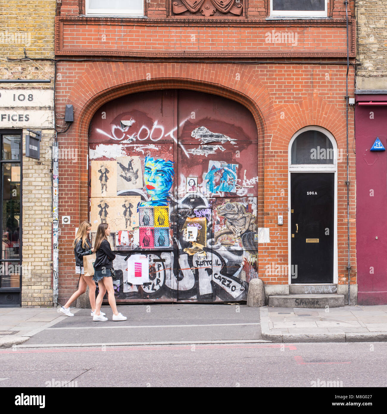 La gente a piedi nella parte anteriore di un vecchio sportello coperto in adesivi e arte di strada murales in Commercial Street, Spitalfields, Shoreditch, East London, Regno Unito Foto Stock