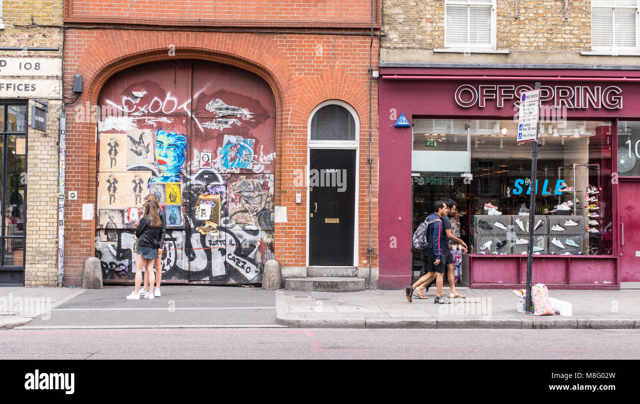 La gente a piedi nella parte anteriore di un vecchio sportello coperto in adesivi e arte di strada murales accanto alla prole store nella strada commerciale, Spitalfields, Shoreditc Foto Stock