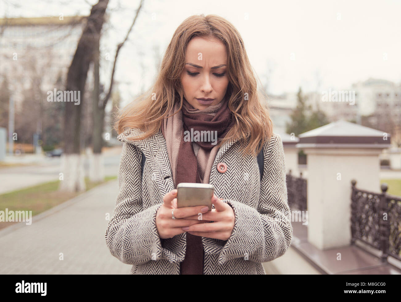 Giovane donna arrabbiato in dubbio indossando cappotto e utilizza lo smartphone sul background urbano. Foto Stock