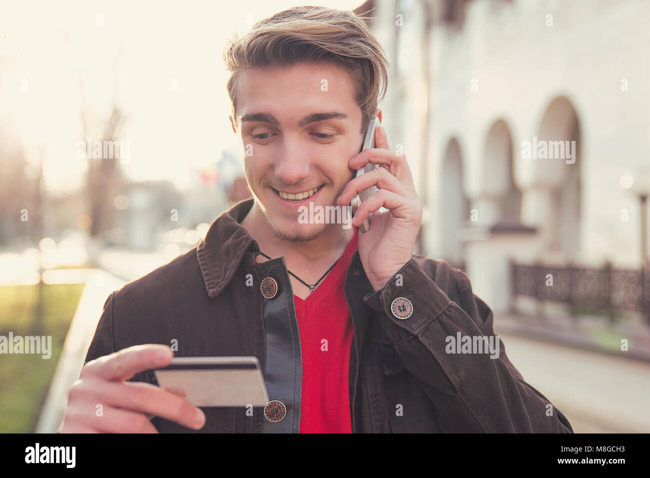 Giovane uomo allegro tenendo la carta di credito e fare ordine tramite chiamata telefonica permanente sulla strada soleggiata. Foto Stock