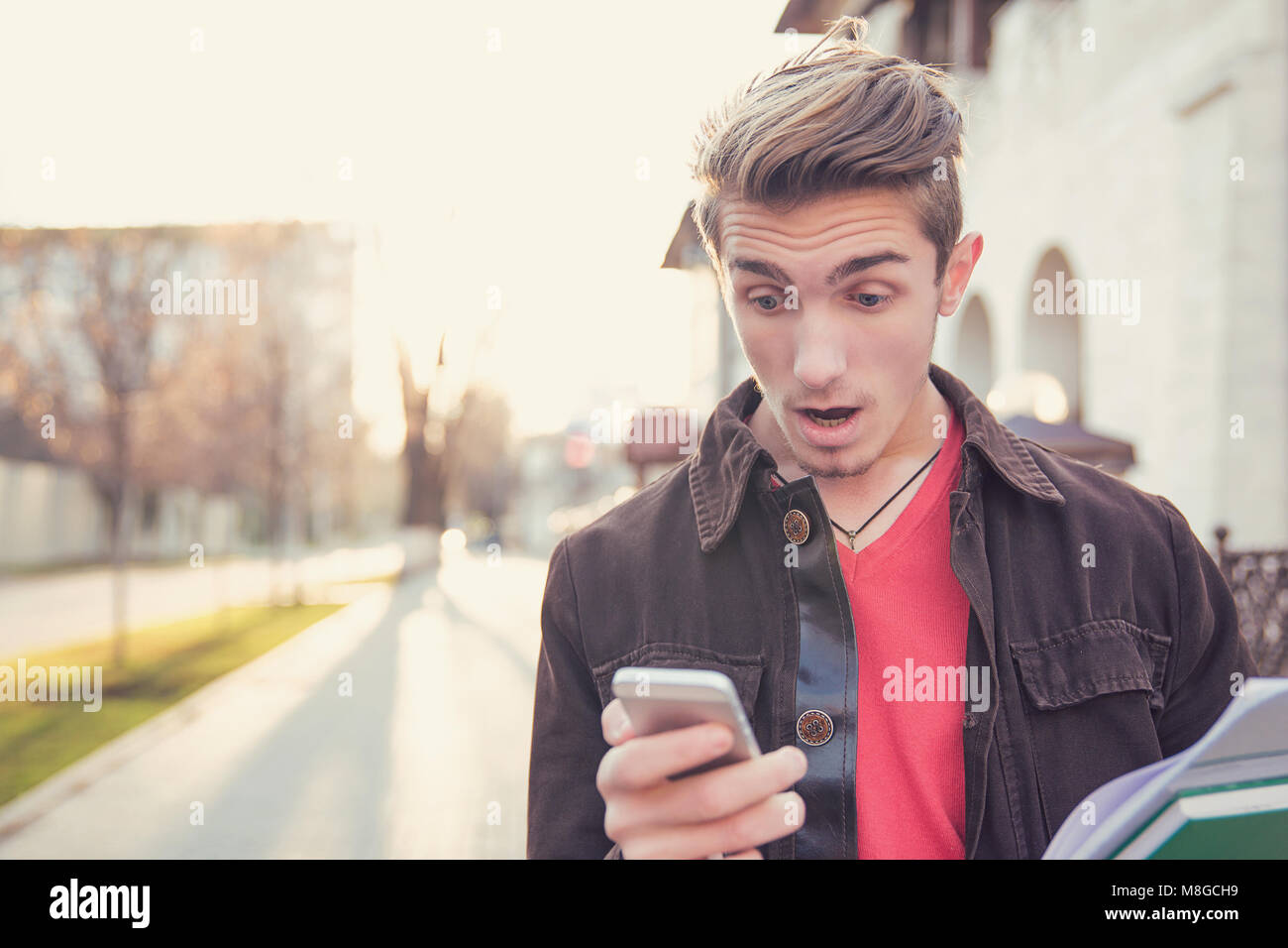 Giovane uomo informale con gli studi controllo news in cerca dello smartphone estremamente sconvolti sul background urbano. Foto Stock