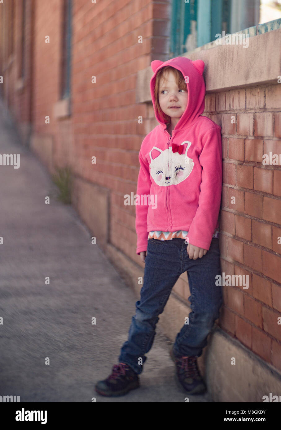 Little Red intitolata girl, appoggiata contro un muro di mattoni, indossando una rosa di Hello Kitty hoodie. Canon EOS 6D, Bausch & Lomb 3 1/4" 82mm f/2.8 Petzval Foto Stock