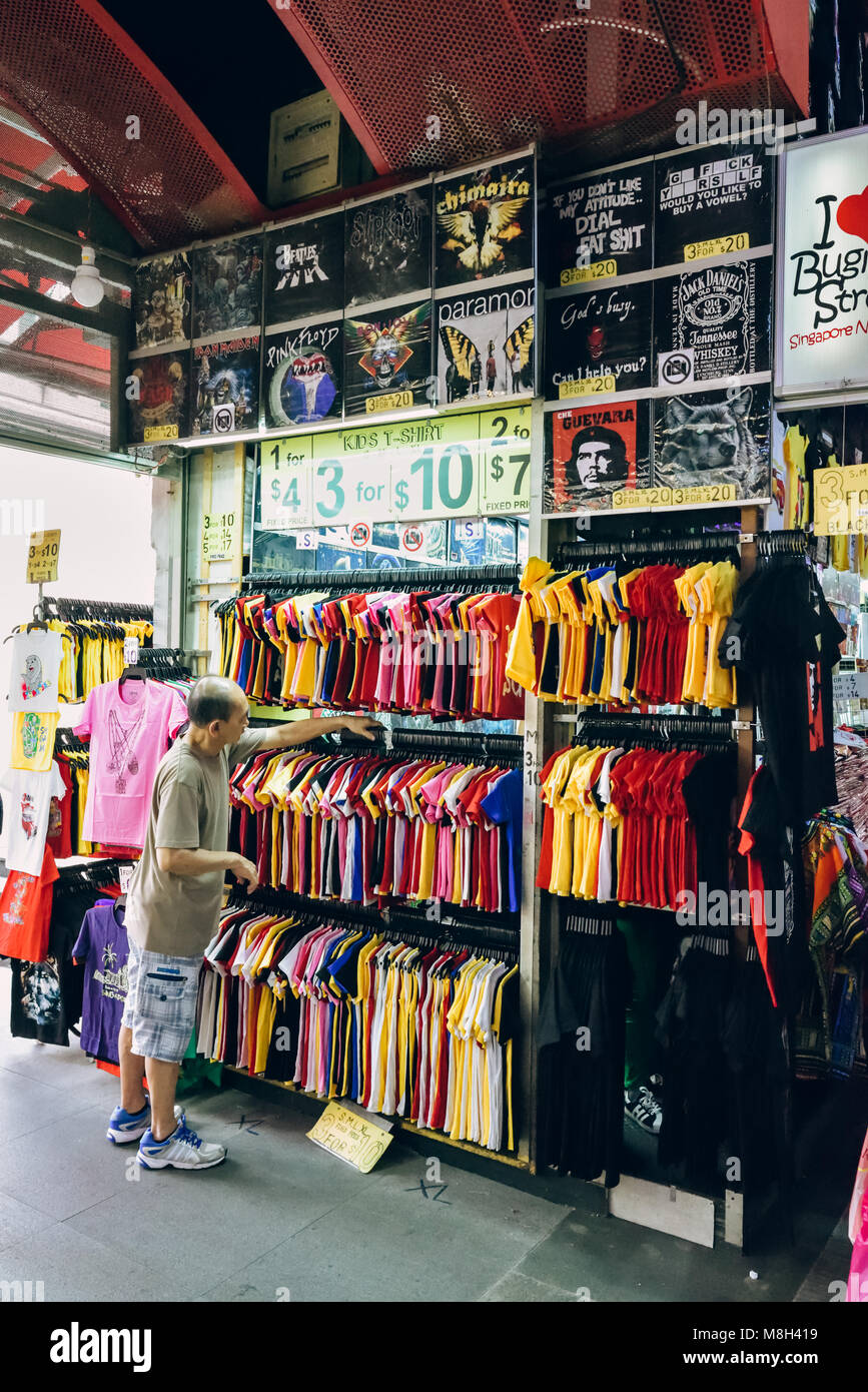 Bugis Street, Singapore. Un negoziante o assistente organizza la sua parete di conveniente appariscente t-shirt per vendita a questo bilancio luogo di shopping. Foto Stock