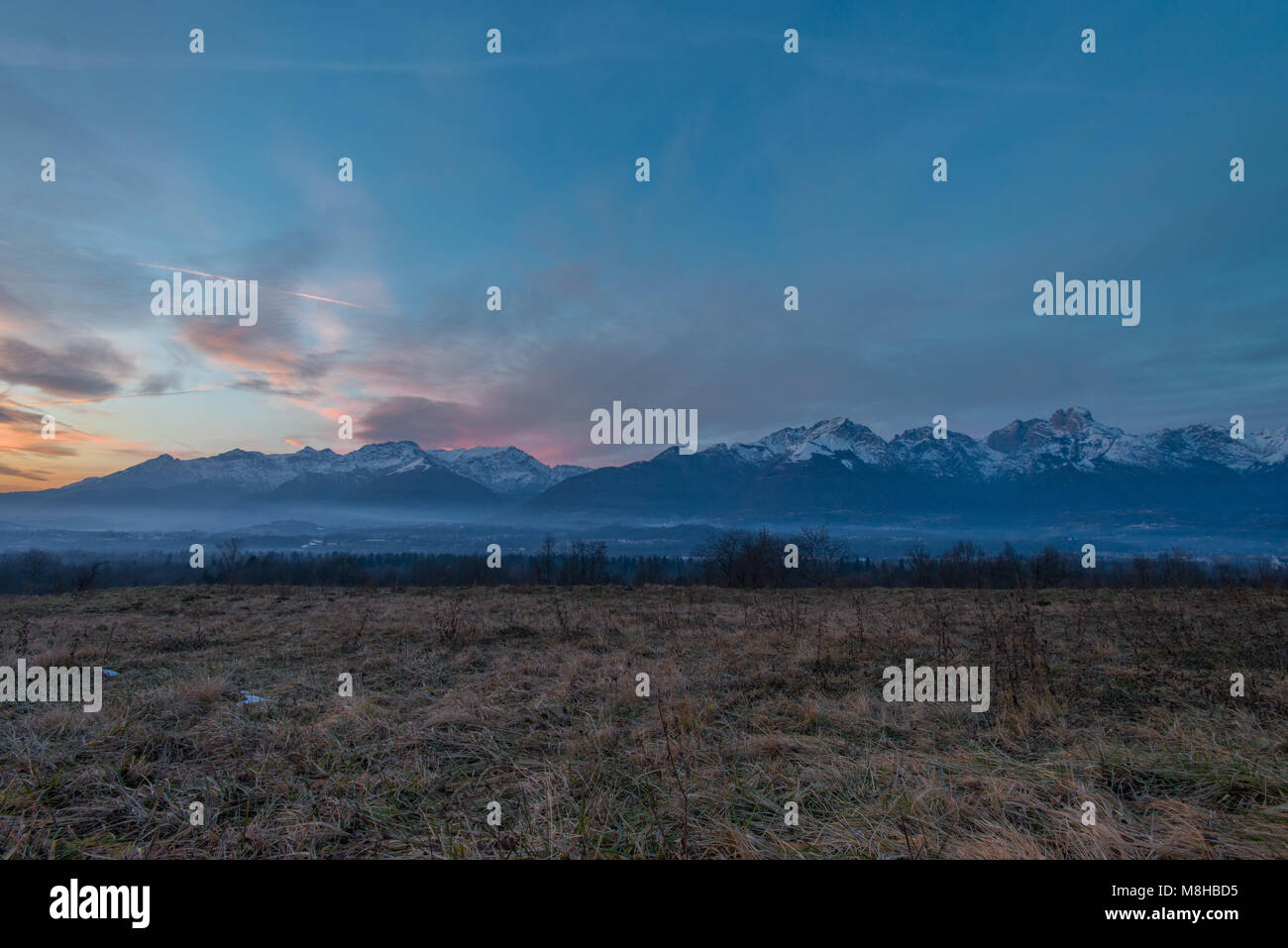 Dipinto il cielo al tramonto, spooky prato ricoperto di haze dà il via ad una imponente catena montuosa con cime ricoperte di neve. Foto Stock