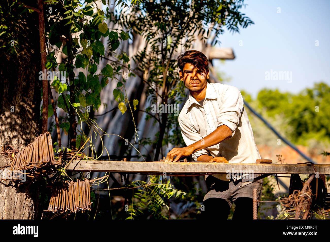 Un bel giovane uomo sta lavorando all' aperto nel villaggio indiano Nipal, piegatura di tondini di ferro durante la bella giornata di sole sotto gli alberi. La luce è golden caldo come Foto Stock