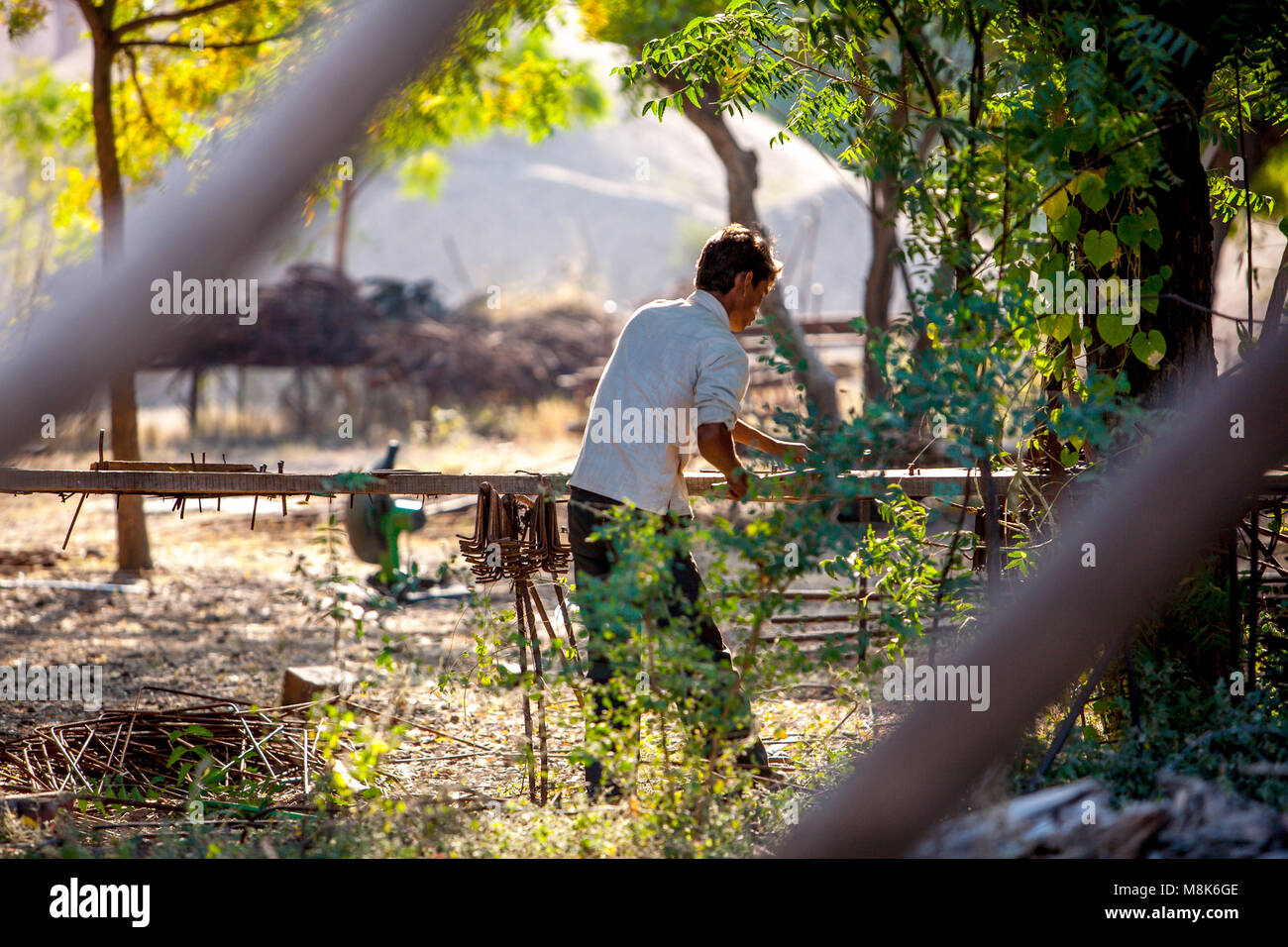 Un bel giovane uomo sta lavorando all' aperto nel villaggio indiano Nipal, piegatura di tondini di ferro durante la bella giornata di sole sotto gli alberi. La luce è golden caldo come Foto Stock