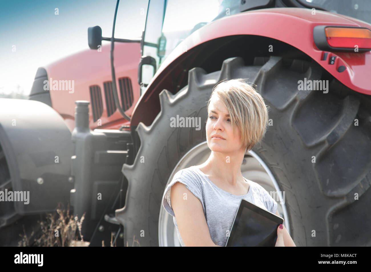Piuttosto giovane donna con la compressa in piedi in campo con il trattore in background Foto Stock