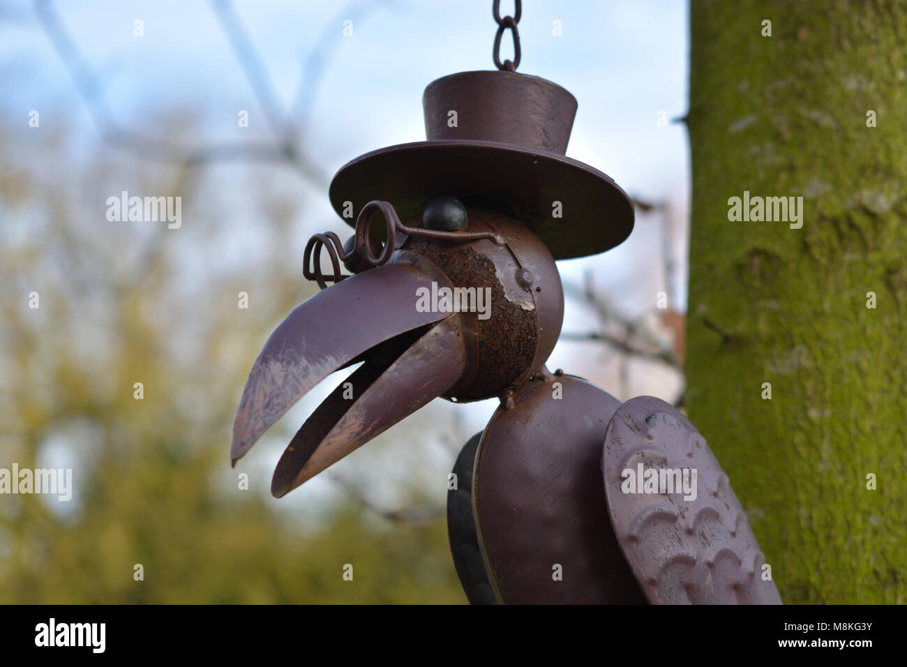 Una stravagante scultura di corvo in metallo con occhiali e un cappello pende da un ramo d'albero. Foto Stock