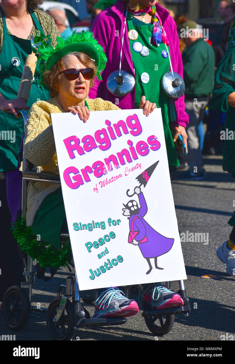 Una donna anziana seduta su una sedia a rotelle con un cartello che dice che infuria nonni di Whatcom County. Indossa un cappello verde e il vestito e si trova nel bel Foto Stock