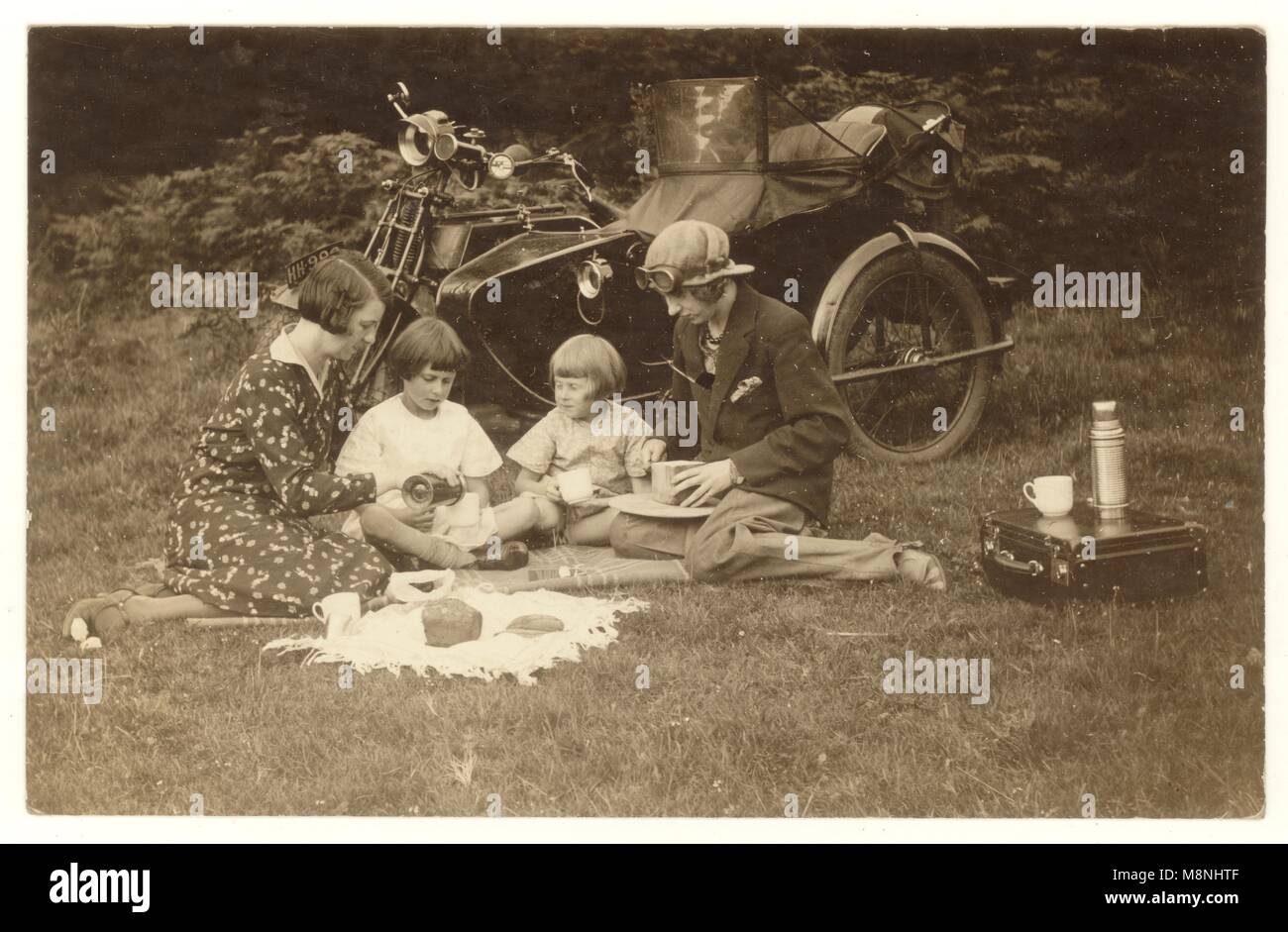 1920's cartolina di nonna con i bambini e la loro madre, avente un picnic accanto a una moto con un side-car, la più giovane donna indossa pantaloni e cappello con occhiali, U.K. Foto Stock