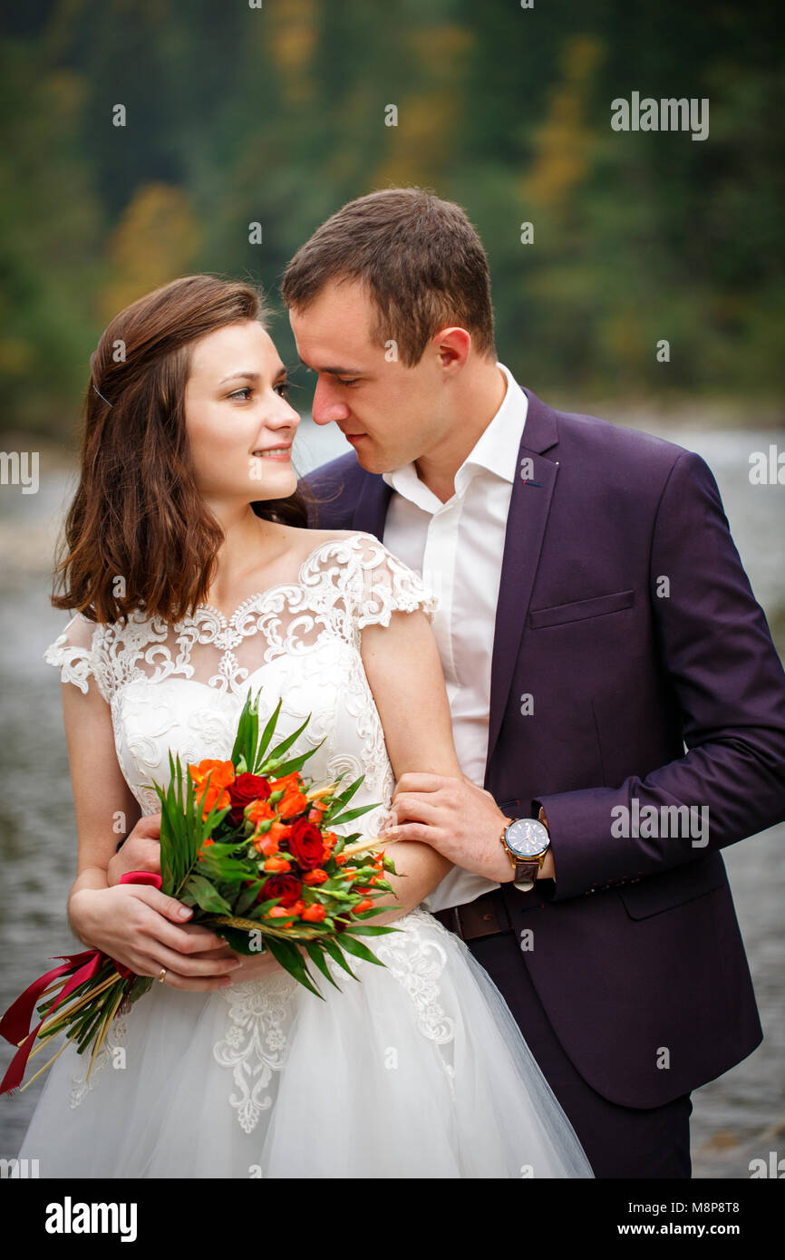 Agli sposi novelli esaminare ogni altra agli occhi, in piedi sulla riva del fiume. Una sposa con un bouquet di fiori dal vivo Foto Stock