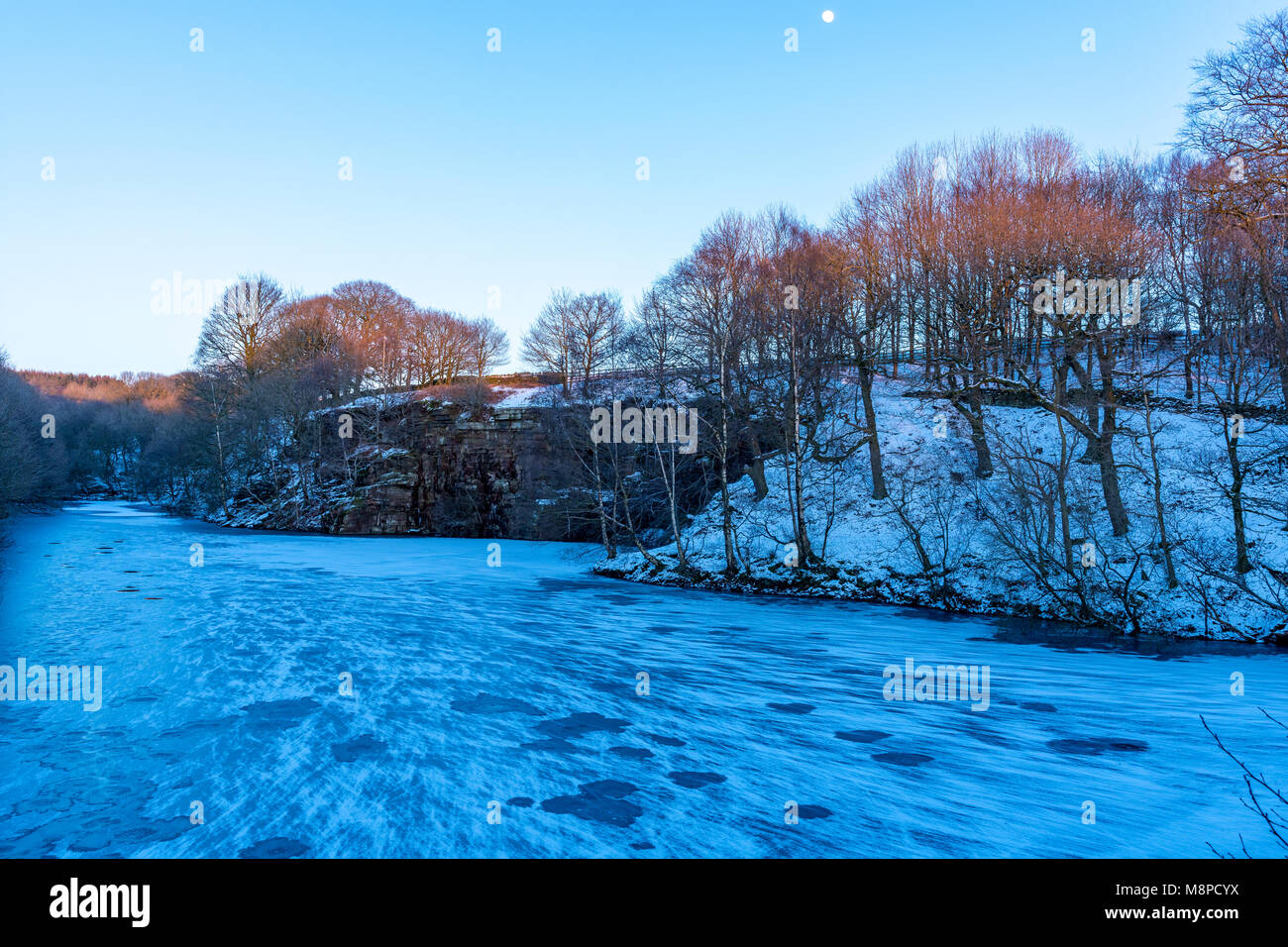 Acque congelate nelle acque del serbatoio Anglezarke Chorley Lancashire Regno Unito come tramonta il sole e la Luna appare nel cielo blu sopra Foto Stock