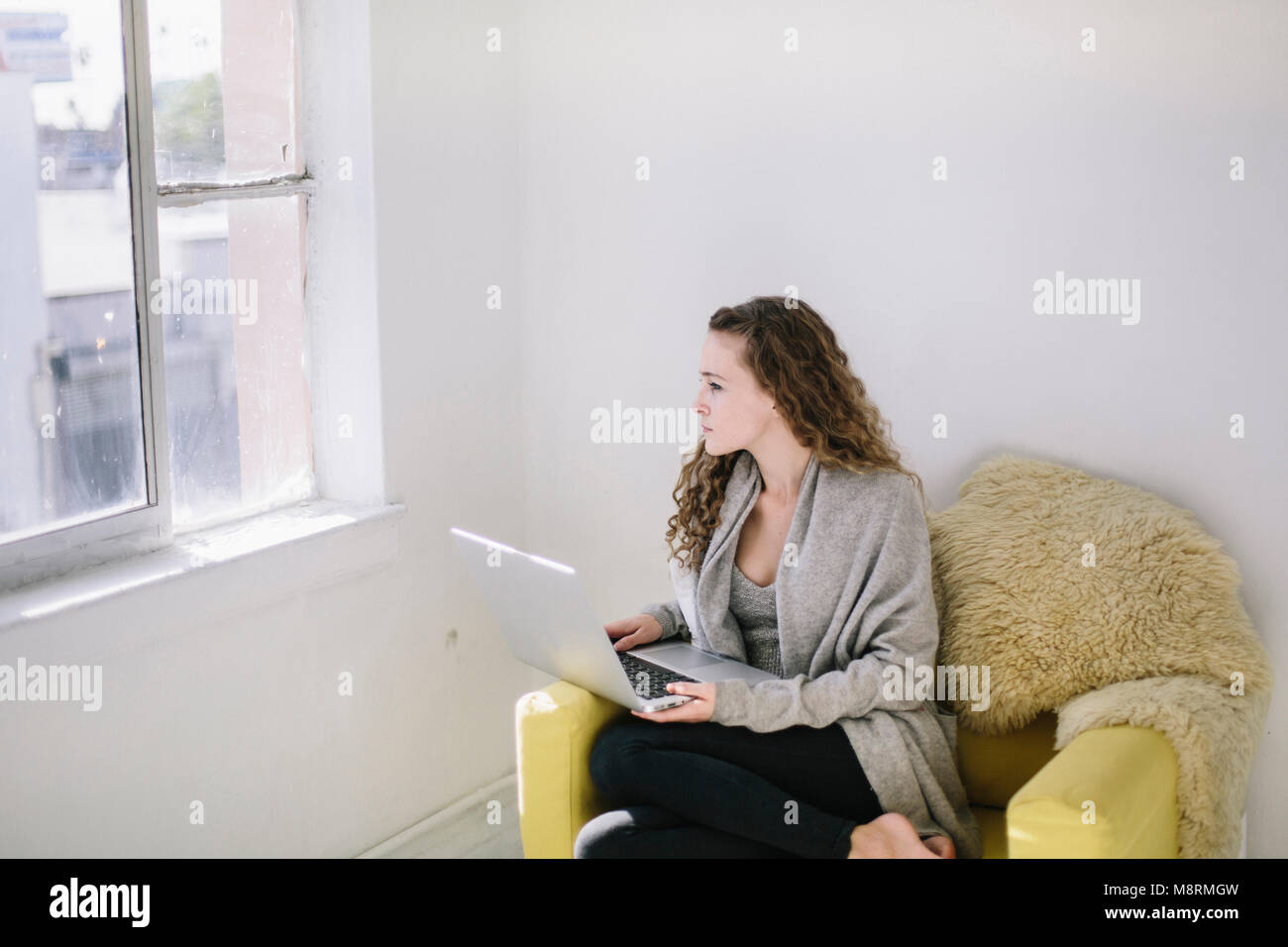 Donna che guarda attraverso la finestra mentre è seduto con il computer portatile su una sedia a casa Foto Stock