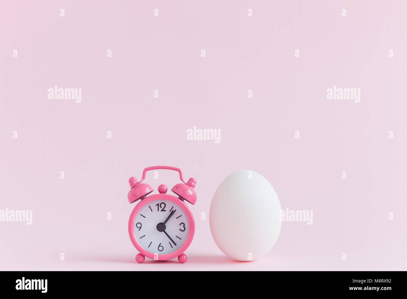 Piccola sveglia e uovo bianco pastello su sfondo rosa. Eastertime la minima nozione. Foto Stock