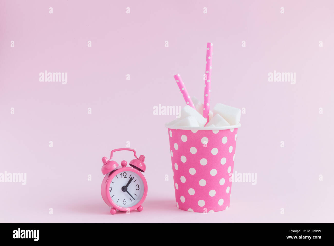 Bicchiere di carta polka design pieno di cubetti di zucchero con acqua potabile cannucce e piccola sveglia pastello su sfondo rosa la minima nozione. Foto Stock