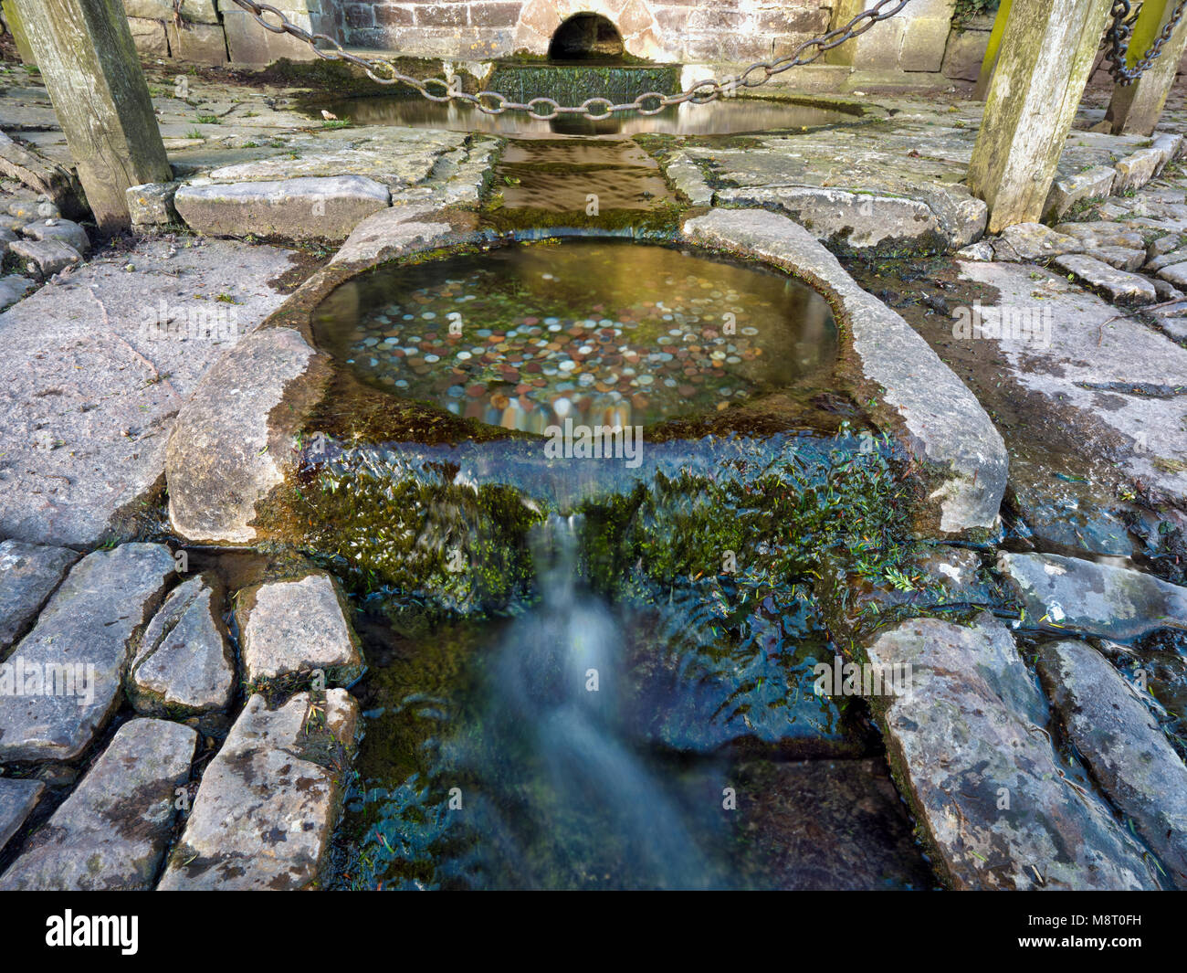 Acqua fresca che scorre da un pozzetto a Tissington villaggio con donazioni di denaro per desideri nel Parco Nazionale di Peak District, Derbyshire, England, Regno Unito Foto Stock