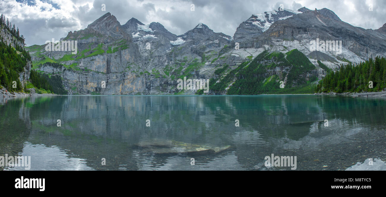 Fantastica vista panoramica del lago Oeschinen, alpine tarn, Alpi Svizzere. Il lago di smeraldo, profonde acque blu, ancora in superficie. Forest, a strapiombo sulle montagne e ghiacciai Foto Stock