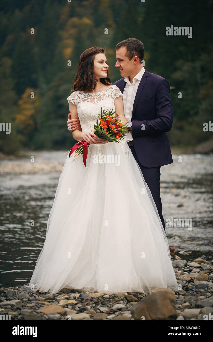 Elegante felice bruna sposa e splendida groom sullo sfondo di un bel fiume in montagna Foto Stock