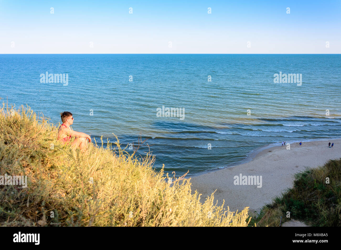 Un giovane uomo in una maglietta rossa si siede su una collina sullo sfondo del mare 2018 Foto Stock