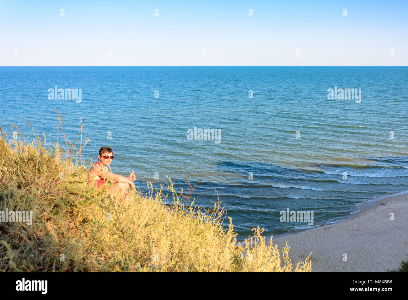 Un giovane uomo in una maglietta rossa si siede su una collina sullo sfondo del mare 2018 Foto Stock