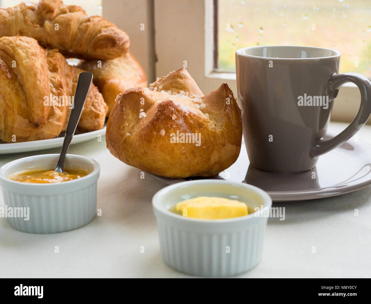 Vista ravvicinata di una brioche, Francese bun , croissant, con burro e marmellata e una tazza di caffè davanti a una finestra in un giorno di pioggia Foto Stock