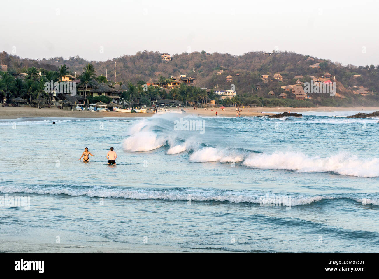 Heavy surf breaks su San Agustinillo spiaggia dove barche da pesca sono spiaggiata & Riva è rivestito con alberi di palma e palapa modeste sistemazioni coperto Foto Stock