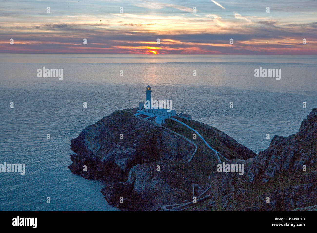 Il sole tramonta dietro il South Stack Lighthouse ad Anglesey, nel Galles del Nord, mentre le condizioni ghiacciate portate dalla 'Bestia dei pini dall'Oriente' sono destinate ad aggrapparsi ancora un giorno prima che la Gran Bretagna possa vedere il ritorno della primavera. Foto Stock