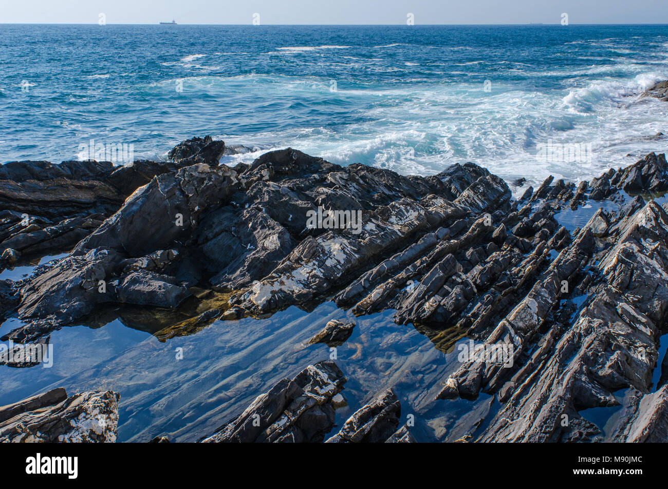 Un mare azzurro acqua intrappolata nelle pietre presso la spiaggia rocciosa di Genova Nervi di fronte al vasto mare mediterraneo con una solitaria lontana nave presso Horizon Foto Stock