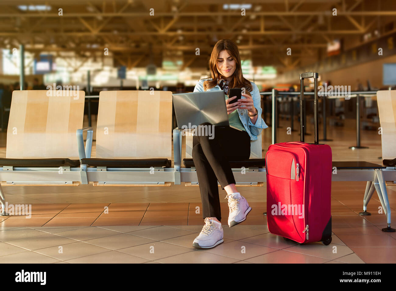 Aeroporto femmina giovane passeggero su smart phone e il computer portatile seduti nella sala terminale durante l'attesa per il suo volo Foto Stock