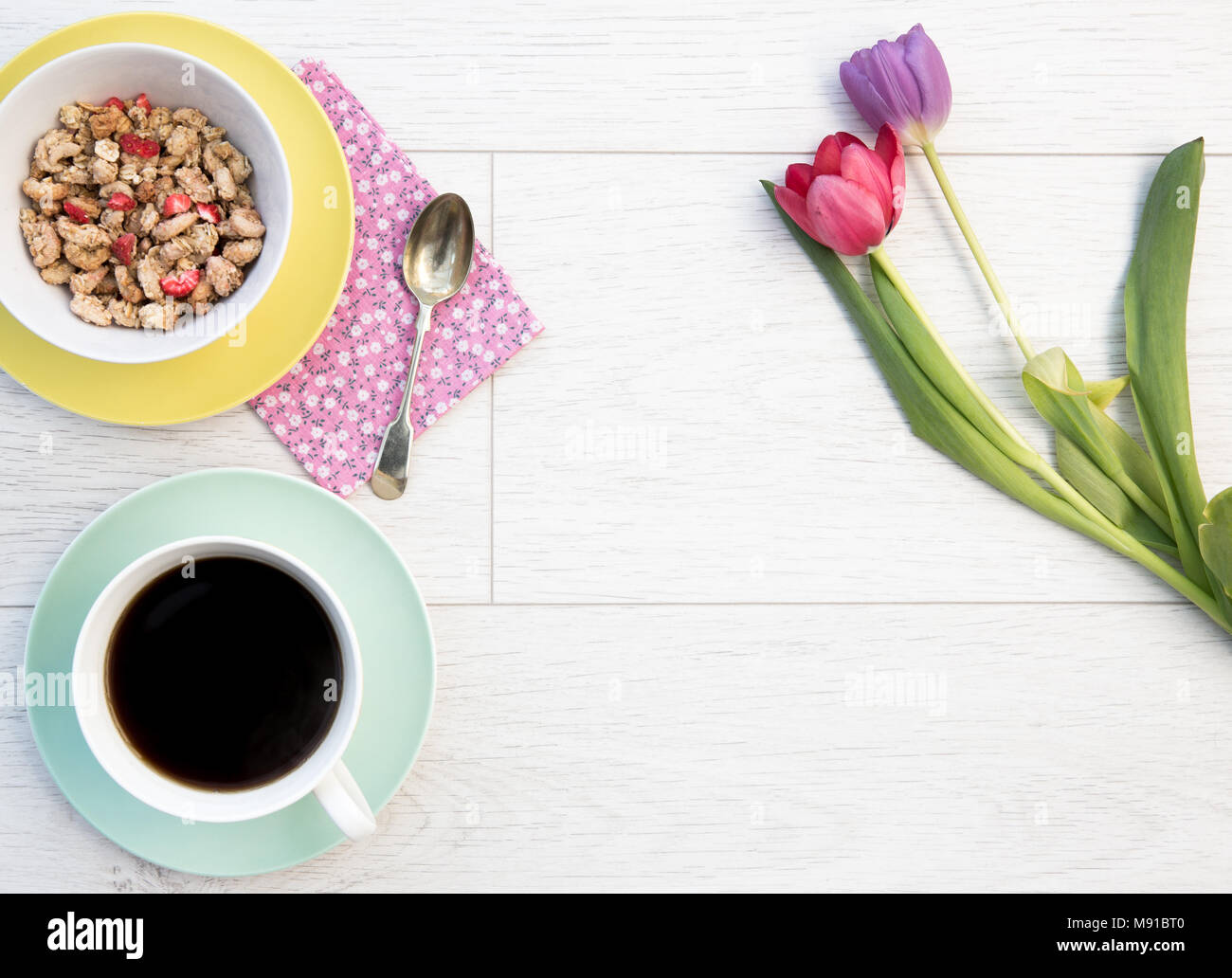 Guardando verso il basso a partire da sopra su un bianco, tavolo in legno top con una colazione femminile di muesli e caffè lungo con i tulipani, fiori e copia di spazio. Foto Stock