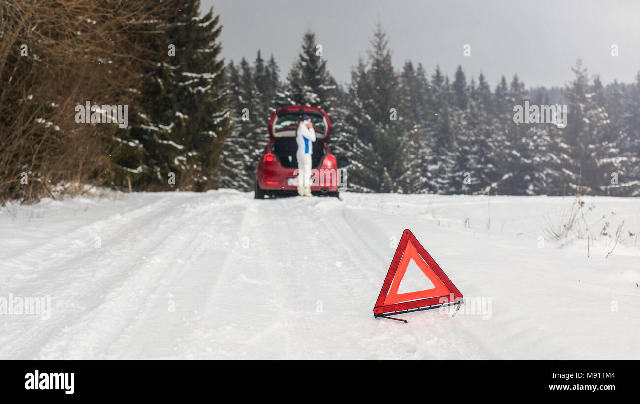 Brillante triangolo rosso su una strada innevata avvertenza altri piloti con la donna in piedi accanto a Broken auto in foresta, chiamando il servizio di riparazione sul suo telefono cellulare Foto Stock