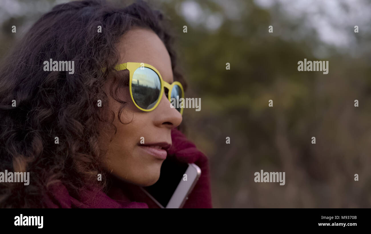 Considerato razza mista donna in giallo occhiali da sole parlando sullo smartphone, close-up Foto Stock
